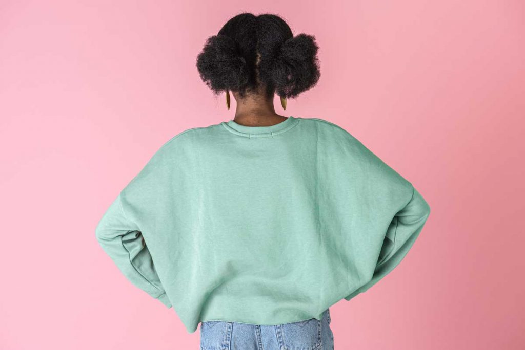 African American woman wearing pastel green sweater with baby blue jeans standing against a pink background