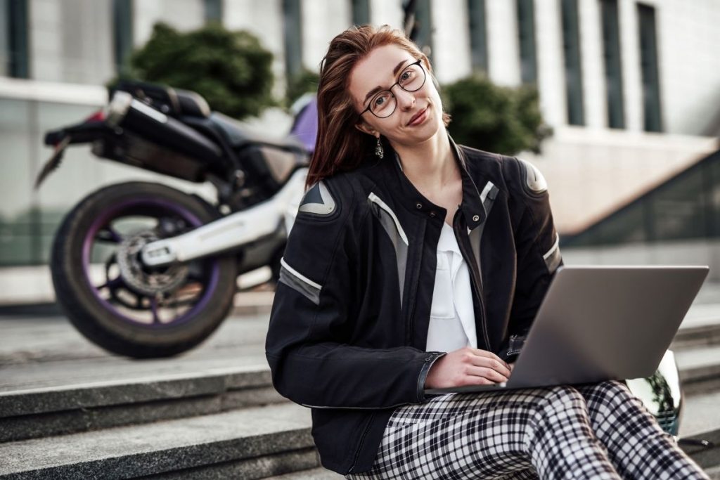 girl working on laptop while sitting on steps wearing black and white plaid pants with a white blouse and black and white motorcycle jacket, purple motorcycle in background