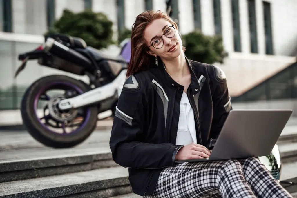 girl working on laptop while sitting on steps wearing black and white plaid pants with a white blouse and black and white motorcycle jacket, purple motorcycle in background