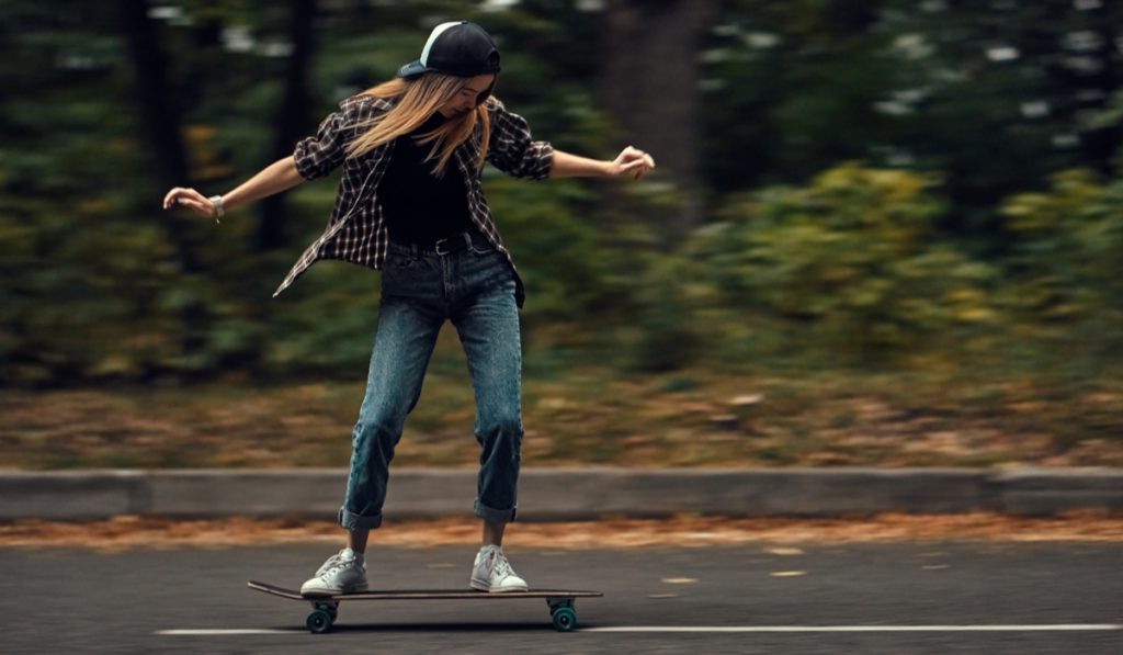 A girl skateboarder in a cap and checkered shirt rides a skateboard on the road in the forest.