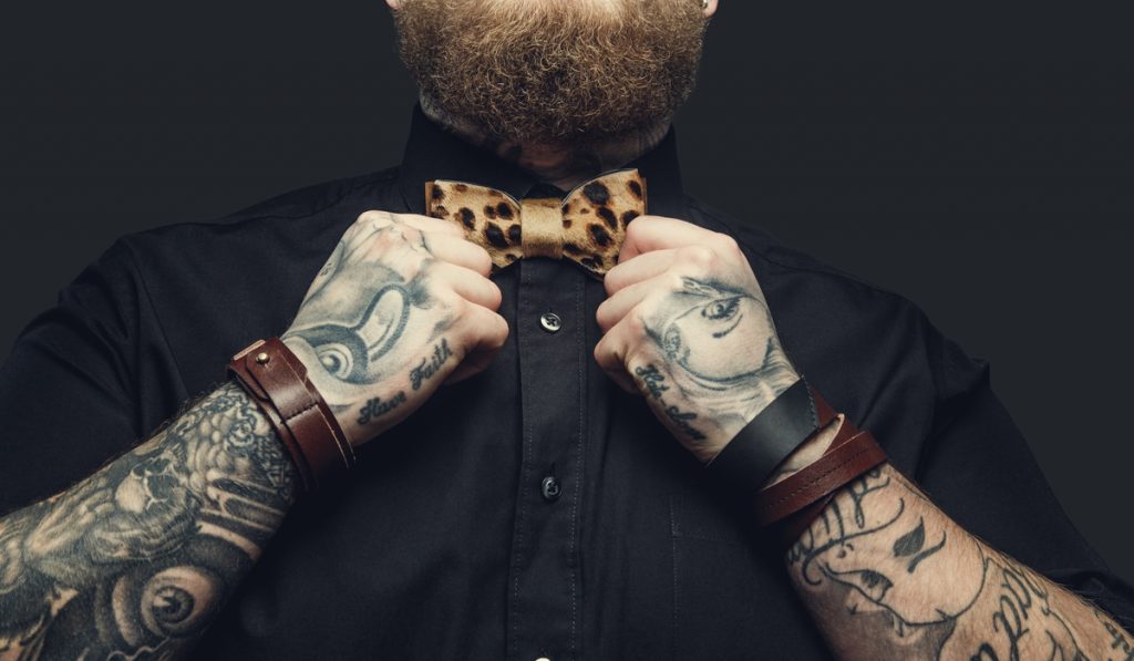 Bearded male with tattooes on his arms in black shirt and brown bow tie.