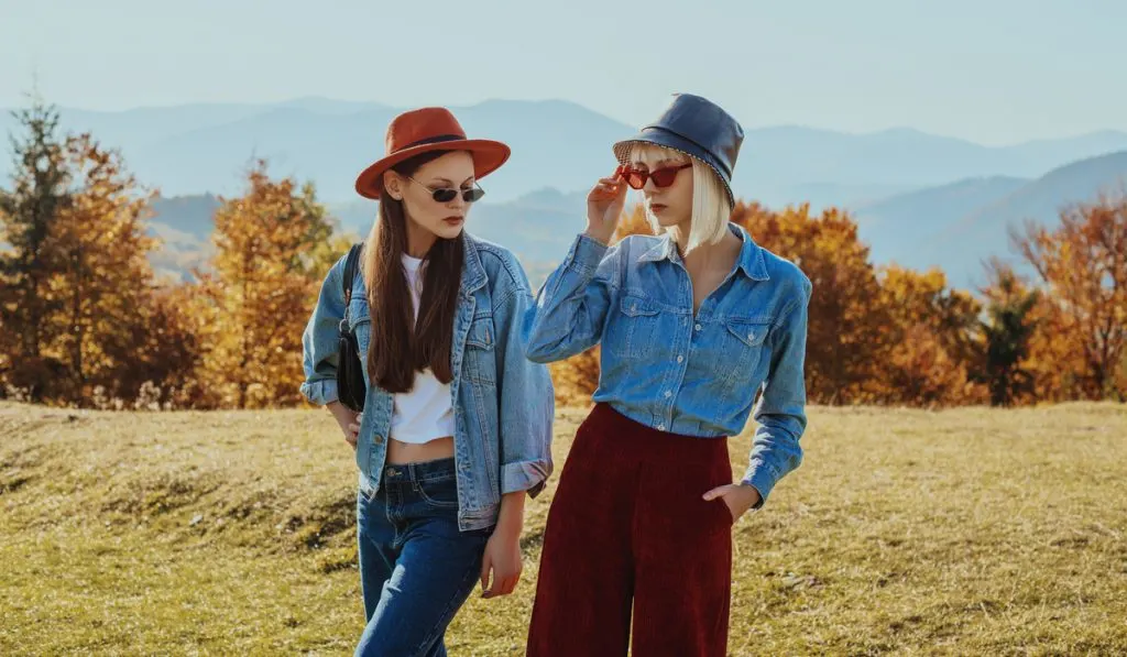 Outdoor autumn fashion portrait of two women, models wearing trendy outfits with hats